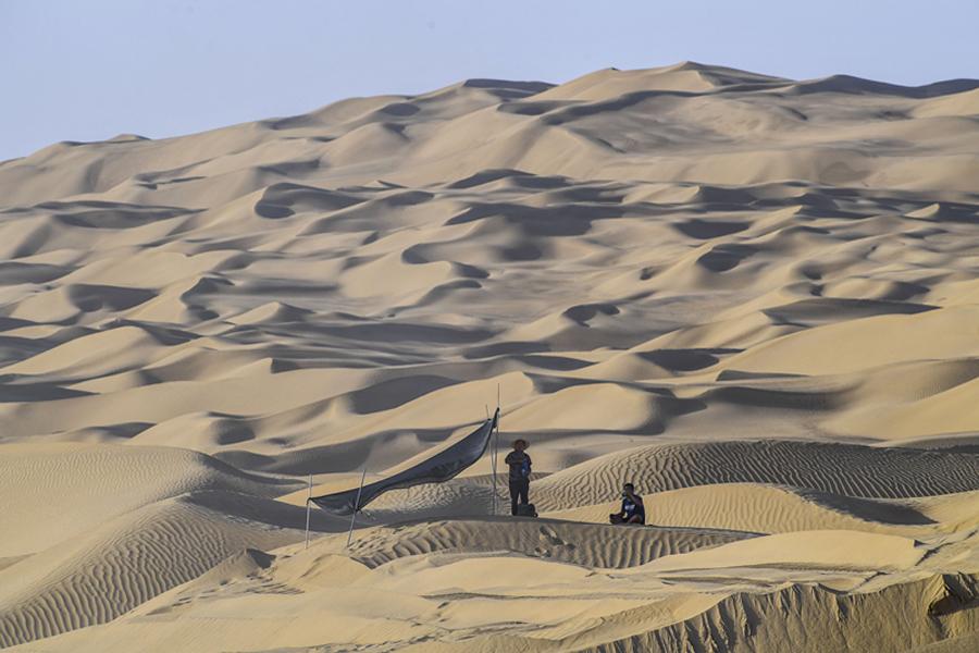 On a sand dune, two workers have a break near a temporary mat shelter. (Photo/Xinhua)
Workers are building only the third paved artery through the Taklimakan Desert to help people and products move more freely
Five bulldozers were working on the side of a 9-meter-high dune. In one of them sat Zhang Xiaodong, his cab swirling with sand. Fine dust had collected in the lines of his sweaty face.
The construction team is building a new road through the Taklimakan Desert, the largest desert in China, covering 337,000 square kilometers in the Xinjiang Uygur autonomous region.
When it is finished, the road will connect the counties of Qiemo in the north and Yuli in the south, becoming only the third paved artery through the vast expanse known as the \
