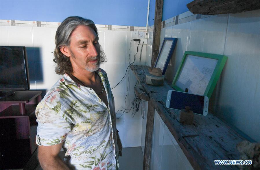 Canadian Craig Anderson shows a mobile phone stand made from driftwood at the eco-sustainability service center of the West Island in Sanya, south China\'s Hainan Province, Sept. 19, 2018. Anderson arrived in February at the West Island inhabited by around 4,000 people. Locals regarded Anderson as a scavenger when he first arrived, as he wandered around the beach almost every day, picking up driftwood, plastic bottles, shattered glasses, and rusty pieces of iron. As time went by, the locals realized that Anderson is an artist who devotes himself in environment protection art design. He profits the recyclable and regenerative materials in making art works. (Xinhua/Yang Guanyu)