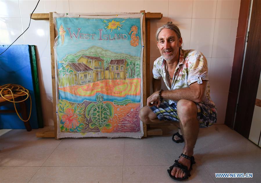 Canadian Craig Anderson poses for a photo beside his painting at the eco-sustainability service center of the West Island in Sanya, south China\'s Hainan Province, Sept. 19, 2018. Anderson arrived in February at the West Island inhabited by around 4,000 people. Locals regarded Anderson as a scavenger when he first arrived, as he wandered around the beach almost every day, picking up driftwood, plastic bottles, shattered glasses, and rusty pieces of iron. As time went by, the locals realized that Anderson is an artist who devotes himself in environment protection art design. He profits the recyclable and regenerative materials in making art works.(Xinhua/Yang Guanyu)