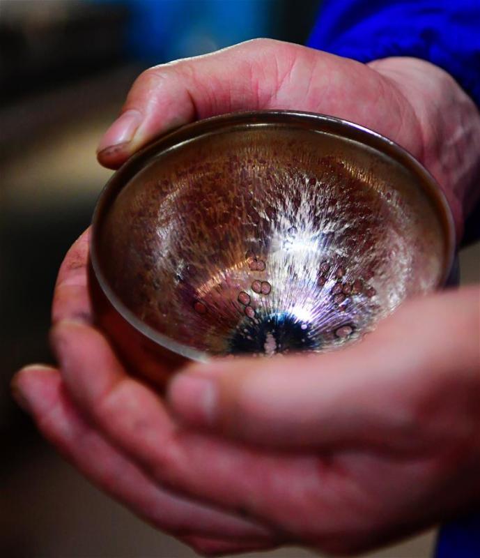 Wu Zhoufu, inheritor of the workmanship of Jianzhan teaware, shows a piece of porcelain at his workshop in Jianyang District of Nanping City, south China\'s Fujian Province, March 28, 2019. Jianzhan teaware, a well-known Chinese porcelain originated in Jianyang, dates back to more than 1,000 years ago in Song dynasty (960-1279). It was the best teaware for scholars and literati to use during that time in tea competitions. Jianzhan teawares are known for their variability. Glazes can range in color from dark plum to yellow, green, and blue. During the heating and cooling processes, iron element in the clay can migrate within the glaze to form surface crystals in rich and glossy colors, as in the \
